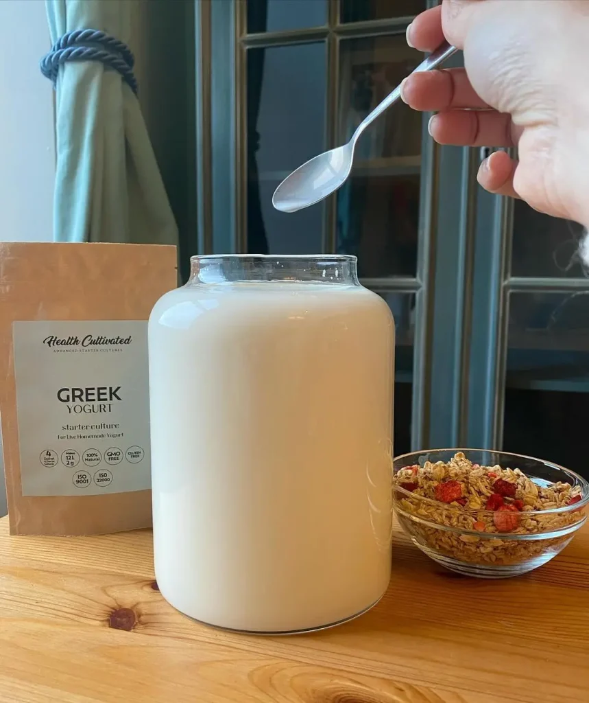 A jar of freshly made Greek yogurt from Health Cultivated sits on a wooden table next to a packet of Greek yogurt starter culture and a bowl of granola with dried fruit.