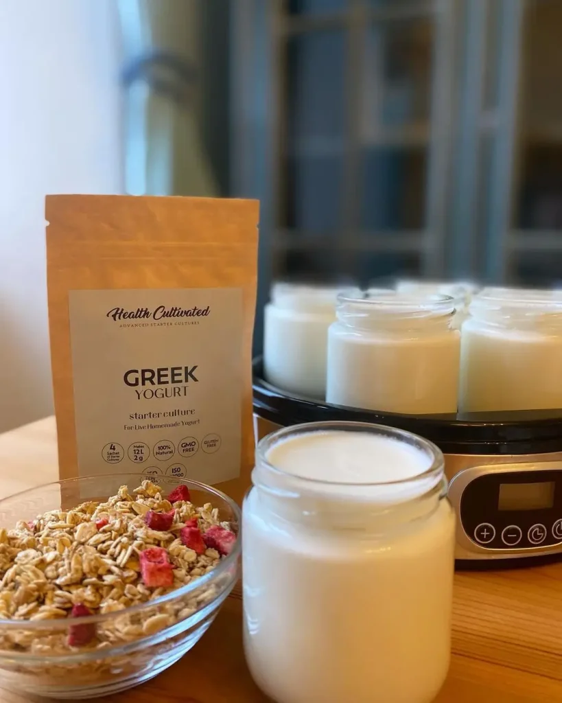 A jar of freshly made Greek yogurt sits on a wooden table next to a packet of Health Cultivated Greek yogurt starter culture and a bowl of granola with dried fruit. In the background, several jars of yogurt are being prepared in a yogurt maker. Soft light filters through a window, creating a cozy kitchen scene.