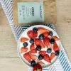 A bowl of Greek yogurt topped with fresh sliced strawberries and blueberries, placed on a striped cloth, next to a brown package "Greek Yogurt starter culture."