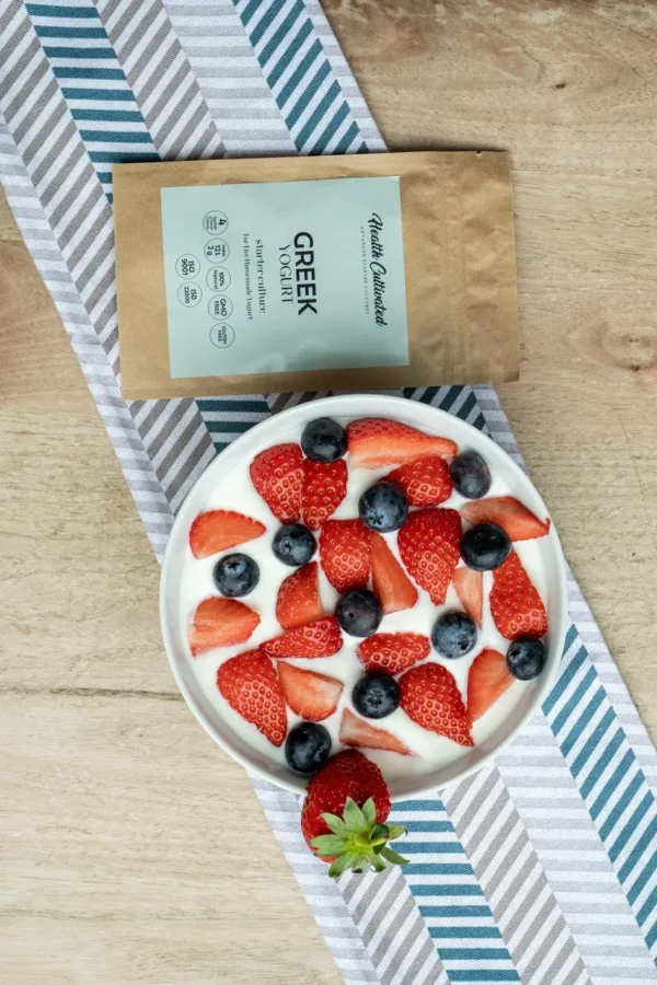 A bowl of Greek yogurt topped with fresh sliced strawberries and blueberries, placed on a striped cloth, next to a brown package "Greek Yogurt starter culture."